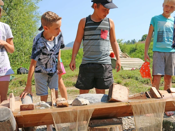 Children are standing in front of a wooden trough; water is flowing over it