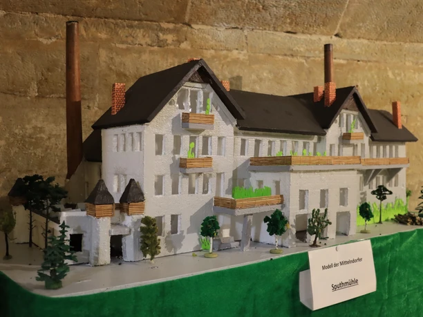 Miniatuurmodel van een wit gebouw met een donker dak en schoorstenen, omringd door kleine bomen, uitgestald op een groene tafel.