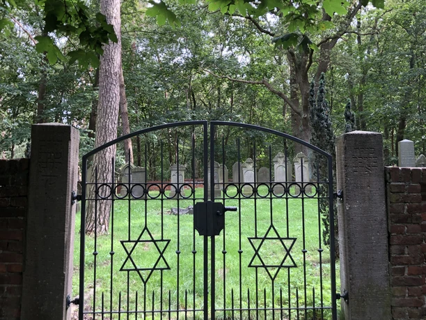 Jüdischer Friedhof Historischer jüdischer Friedhof mit schwarzem Eisentor, verzierten Davidsternen und grünem Baumhintergrund.