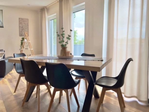 Esszimmer Ferienwohnung Well-Being Esszimmer mit Holzfußboden, modernem Tisch, sechs Stühlen und großen Fenstern mit viel Tageslicht.