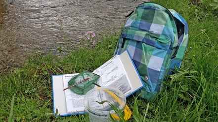 Sac à dos contenant un classeur et du matériel, posé dans un pré au bord d'un ruisseau