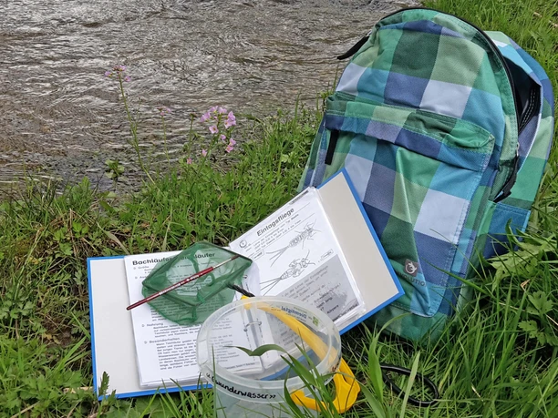 Sac à dos contenant un classeur et du matériel, posé dans un pré au bord d'un ruisseau
