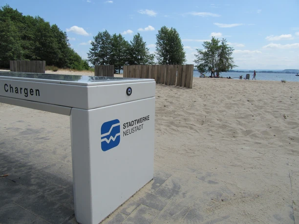 Ladebank Badestrand Weiße Düne Ladebank am Sandstrand mit Bäumen und Wasser im Hintergrund, ideal zum Entspannen und Aufladen.