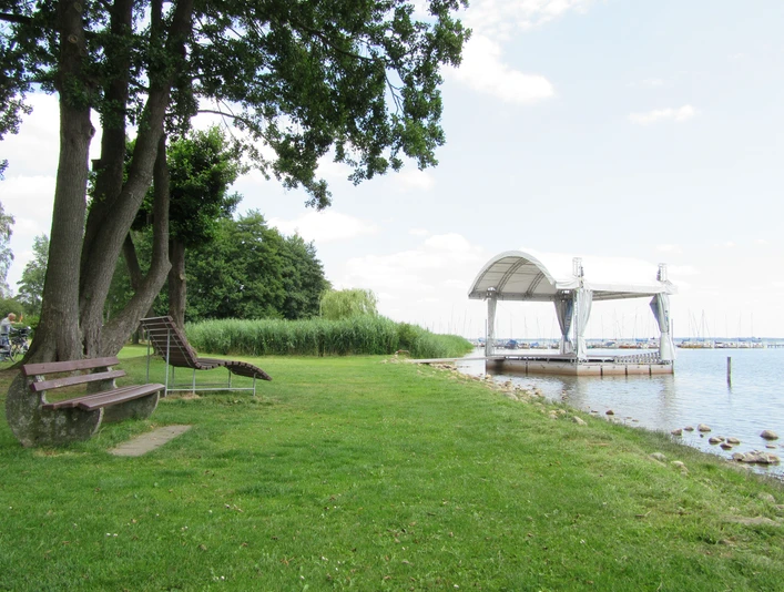 Ein idyllischer Picknickplatz am Ufer mit grüner Wiese, Bäumen und angrenzender Seebrücke.