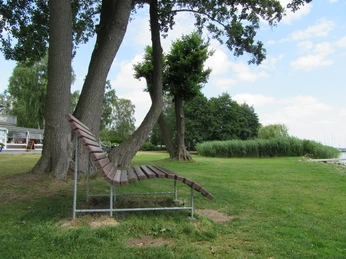 Picknickplatz auf bzw. an der Liegebank Fischerstübchen Holzliege unter Bäumen bietet entspannte Sitzmöglichkeit im Grünen nahe See mit Blick auf Boote.