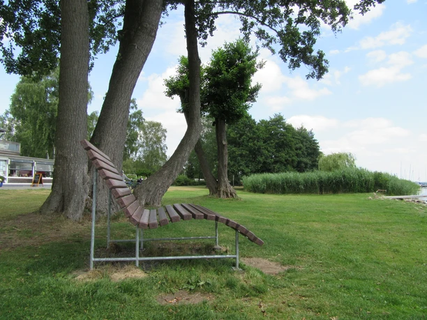 Picknickplatz auf bzw. an der Liegebank Fischerstübchen Holzliege unter Bäumen bietet entspannte Sitzmöglichkeit im Grünen nahe See mit Blick auf Boote.