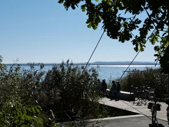 Am Picknickplatz bei der Sehbrücke Mardorf genießen Besucher auf einem Holzsteg den Blick auf das glitzernde Wasser des Steinhuder Meers, umrahmt von Schilf und Bäumen, während Fahrräder angelehnt sind.
