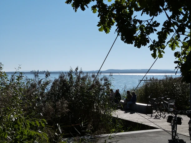 Picknickplatz Sehbrücke Mardorf Am Picknickplatz bei der Sehbrücke Mardorf genießen Besucher auf einem Holzsteg den Blick auf das glitzernde Wasser des Steinhuder Meers, umrahmt von Schilf und Bäumen, während Fahrräder angelehnt sind.