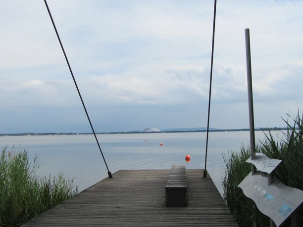 Picknickplatz Sehbrücke Mardorf Ein Holzsteg mit Sitzbank führt zu einem ruhigen See, eingerahmt von Binsen und sanftem Himmel.