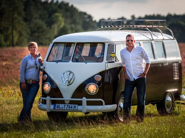 Bulli-Team Paar posiert neben schwarz-weißem VW T1 Bulli in blühender Heidelandschaft.