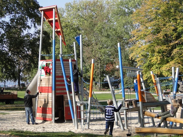 Spielplatz im Park Ein Spielplatz mit Rutsche, Holz- und Kletterstrukturen, Kinder beim Spielen unter Bäumen im Park.
