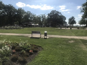 Park mit Blick auf Sonnenuhr Grüne Parklandschaft mit Bank, Weg und Sonnenuhr im Hintergrund, umgeben von Bäumen und Blumenbeet.