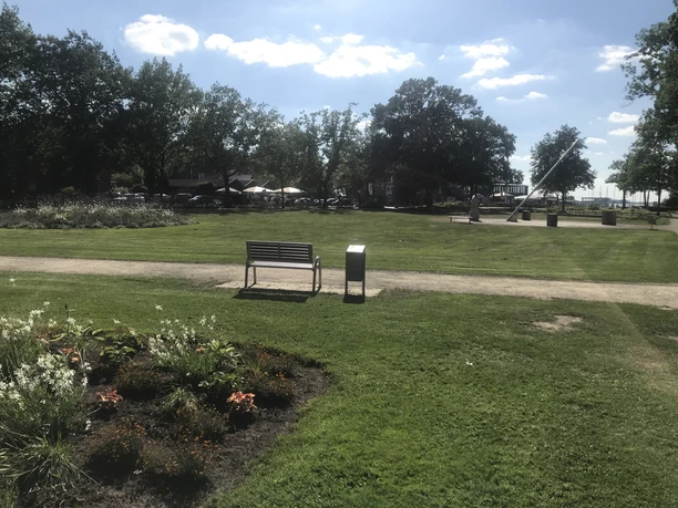 Park mit Blick auf Sonnenuhr Grüne Parklandschaft mit Bank, Weg und Sonnenuhr im Hintergrund, umgeben von Bäumen und Blumenbeet.