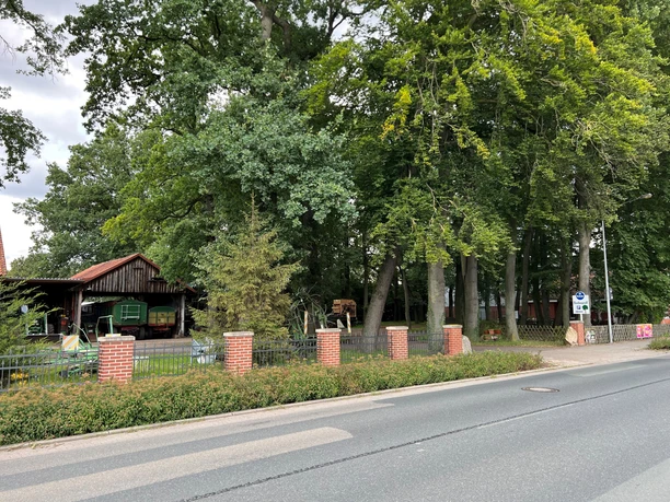 Wohnmobilstellplatz Eystrup Stellplatz mit gepflasterter Zufahrt, umgeben von hohen Bäumen und einer kleinen Holzhütte.