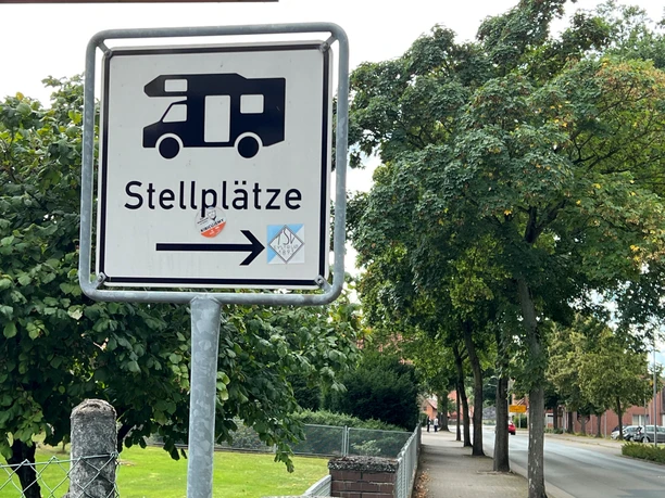 Wohnmobilstellplatz Eystrup Hinweisschild für Wohnmobilstellplätze, flankiert von grünen Bäumen und einem Gehweg am Straßenrand.