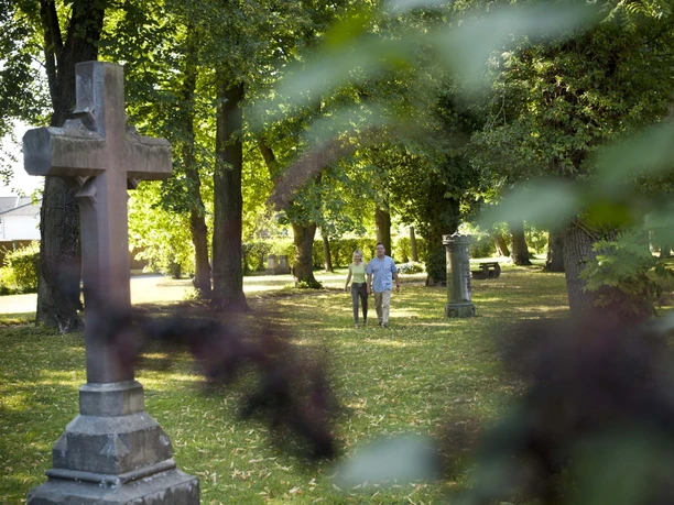 Pärchen auf dem lichtdurchfluteten und mit Bäumen bewachsenem Friedhofswall mit großem Sandsteinkreuz