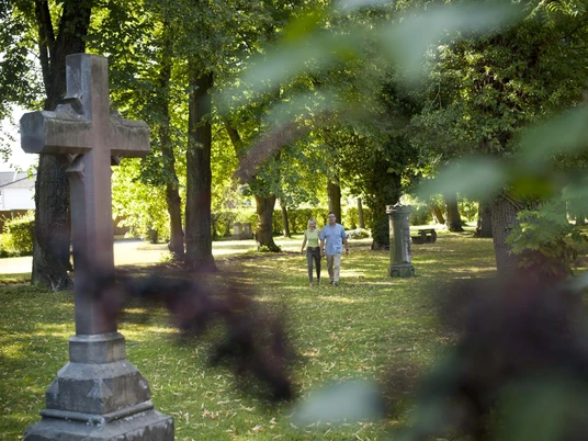 hx-stadtentdecken-andreashub-7 Pärchen auf dem lichtdurchfluteten und mit Bäumen bewachsenem Friedhofswall mit großem Sandsteinkreuz