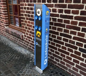 Ladestation Barrierefreie Ladesäule in blau an einer Backsteinmauer mit Logos für Rad und E-Bike-Ladeanschlüsse.