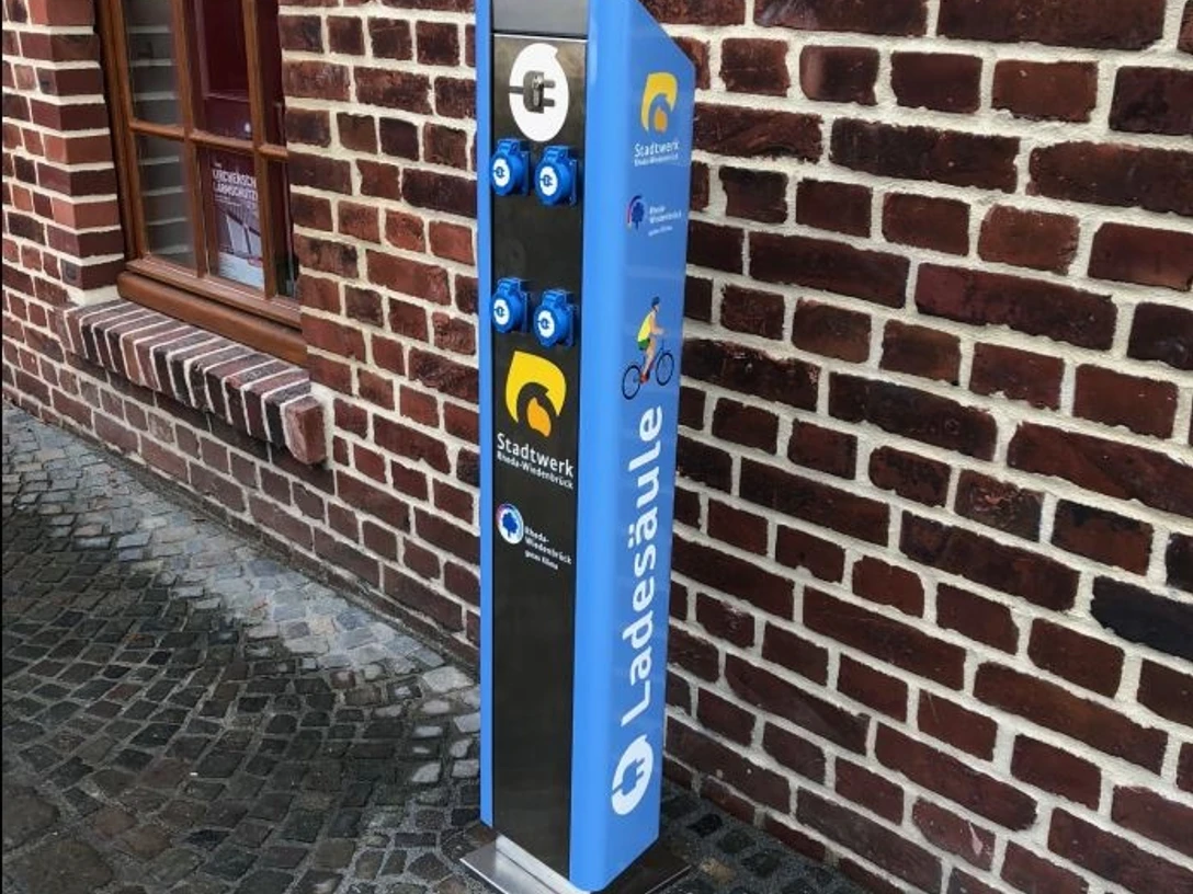 Ladestation Barrierefreie Ladesäule in blau an einer Backsteinmauer mit Logos für Rad und E-Bike-Ladeanschlüsse.