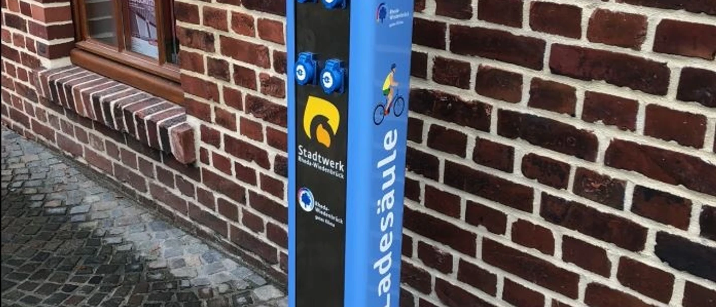 Ladestation Barrierefreie Ladesäule in blau an einer Backsteinmauer mit Logos für Rad und E-Bike-Ladeanschlüsse.