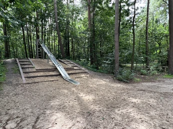 Eine Rutsche im Wald, umgeben von hohen Bäumen, steht auf einem sandigen Spielplatzboden.
