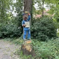 Holzskulptur eines lächelnden Marktschreiers mit blauer Kleidung, auf einem Baumstumpf im Grünen.