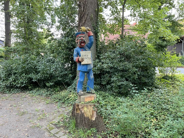 Holzskulptur eines lächelnden Marktschreiers mit blauer Kleidung, auf einem Baumstumpf im Grünen.