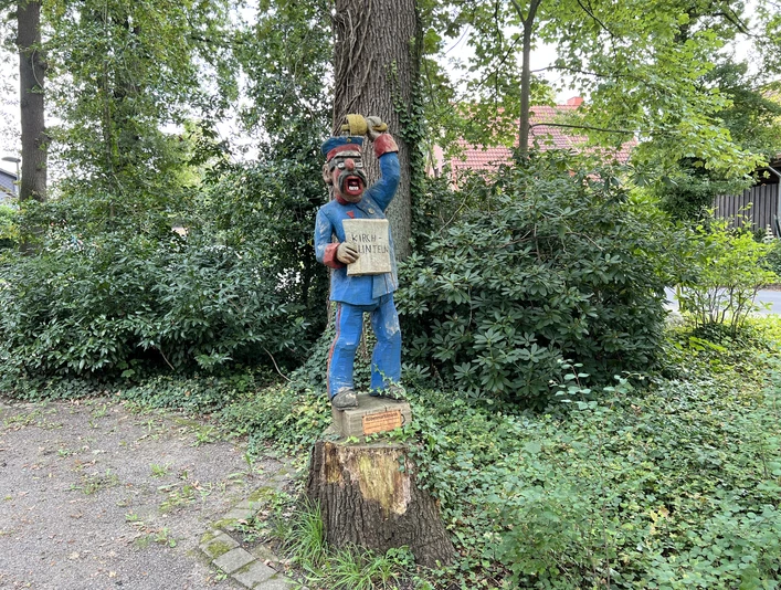 Holzskulptur eines lächelnden Marktschreiers mit blauer Kleidung, auf einem Baumstumpf im Grünen.