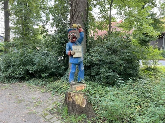 Röbenmarktmann Holzskulptur eines lächelnden Marktschreiers mit blauer Kleidung, auf einem Baumstumpf im Grünen.
