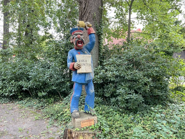 Holzfigur eines Mannes in blauer Uniform mit Tafel "Kirchlinteln" an einem parkähnlichen Ort.
