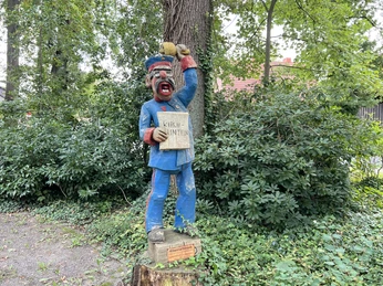 Röbenmarktmann Holzfigur eines Mannes in blauer Uniform mit Tafel "Kirchlinteln" an einem parkähnlichen Ort.