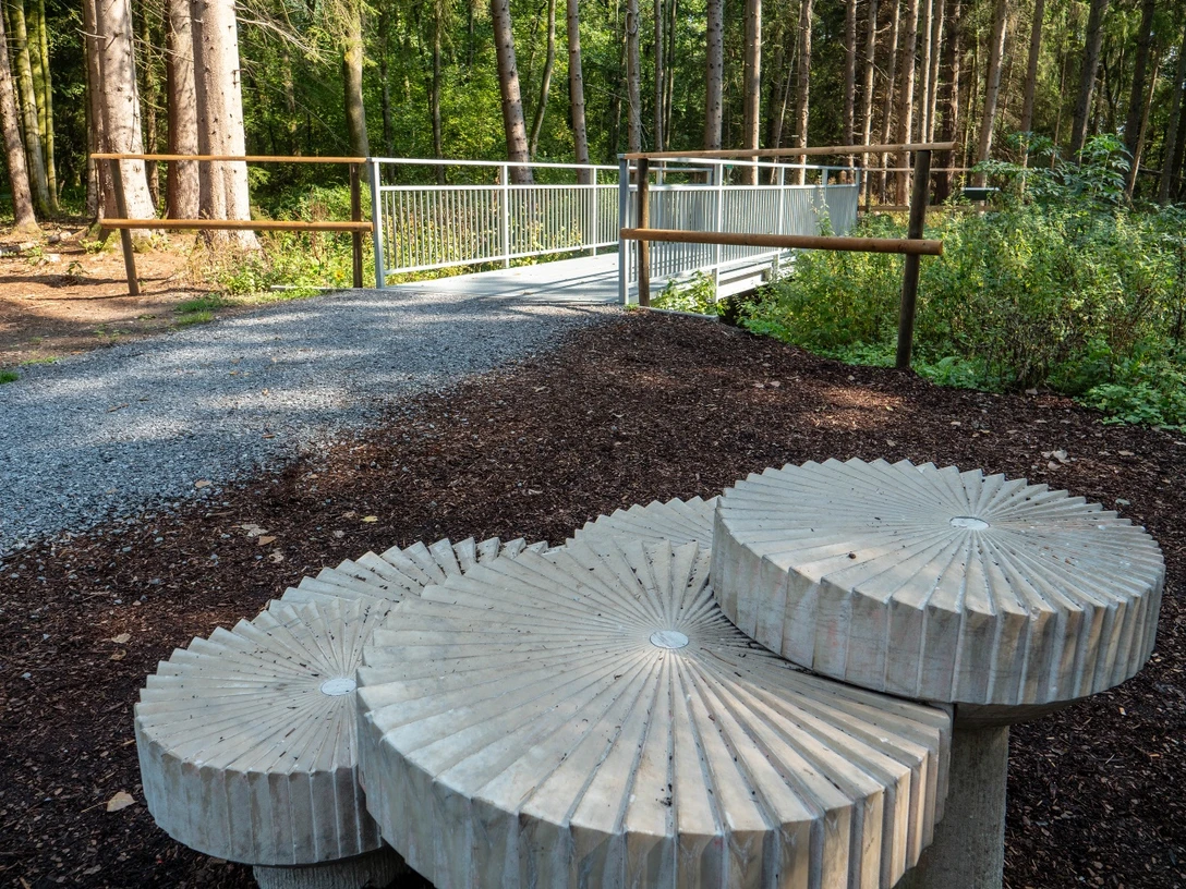 Mühlsteine an der neuen Emsbrücke Drei große, graue Beton-Mühlsteine liegen nahe einer modernen Fußgängerbrücke im Wald.