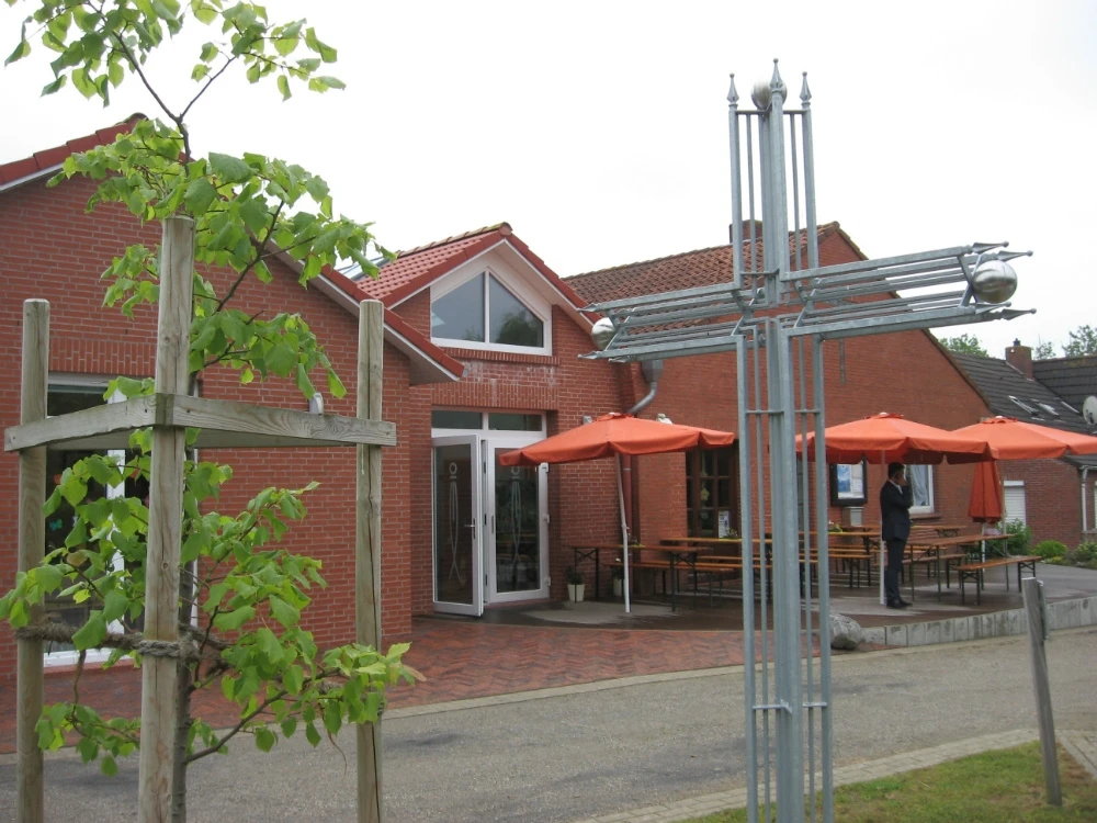 Evangelisch-Freikirchliche Gemeinde Jennelt Backsteingebäude mit großem Metallkreuz und Sitzbereich mit orangefarbenen Sonnenschirmen.