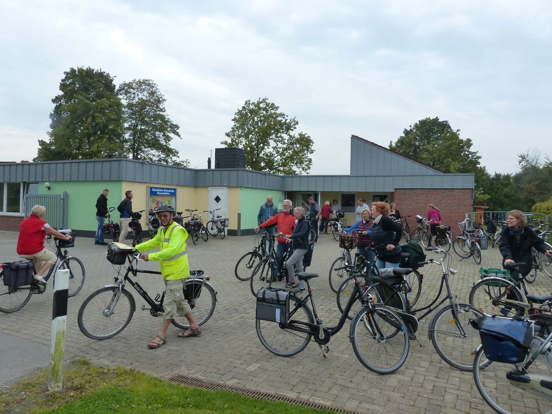 Christliche Gemeinde Krummhörn Gruppe von Radfahrenden vor einem modernen Gemeindehaus mit grün-grauer Fassade und Bäumen im Hintergrund