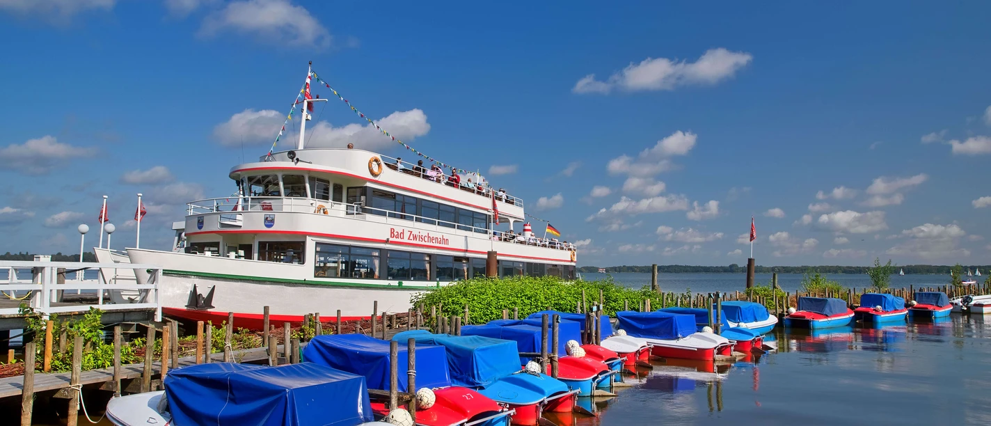 boote-weisse-flotte Ein weißes Ausflugsschiff der Weißen Flotte liegt am Steg, umgeben von farbenfrohen Freizeitbooten.