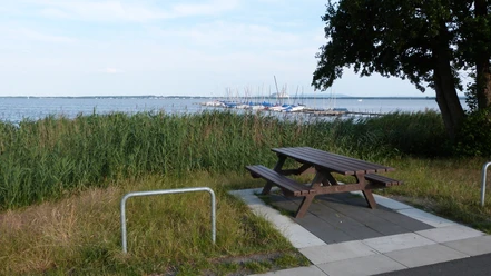 Picknickplatz am Rundweg, mit Blick auf einen See und Boote, umgeben von Schilf und Bäumen.