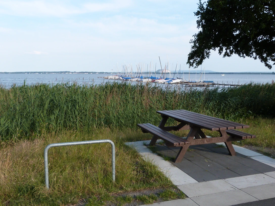 Picknickplatz am Rundweg, mit Blick auf einen See und Boote, umgeben von Schilf und Bäumen.