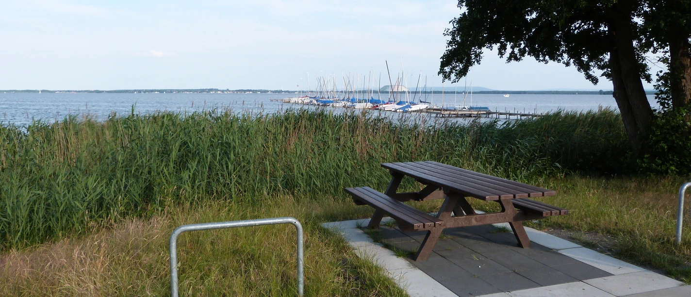 Picknickplatz am Rundweg, mit Blick auf einen See und Boote, umgeben von Schilf und Bäumen.