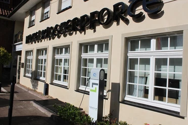 Elektrische Ladestation vor einem beige gestrichenen Bürogebäude mit schwarzen Schriftzügen und Fenstern.