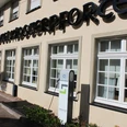 Elektrische Ladestation vor einem beige gestrichenen Bürogebäude mit schwarzen Schriftzügen und Fenstern.