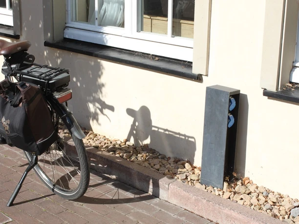 E-Bike-Ladesteckdosen am Klosterhof Ein schwarzes Fahrrad lehnt an einer gelblichen Hauswand neben einem kleinen, grauen Stromkasten.