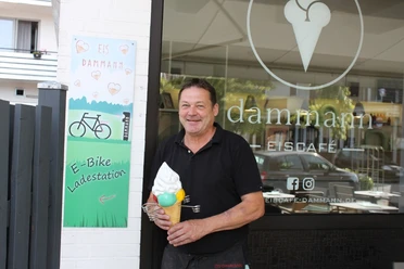 Ein lächelnder Mann hält ein Eis vor dem Eingang eines modernen Eiscafés mit E-Bike-Ladestation.
