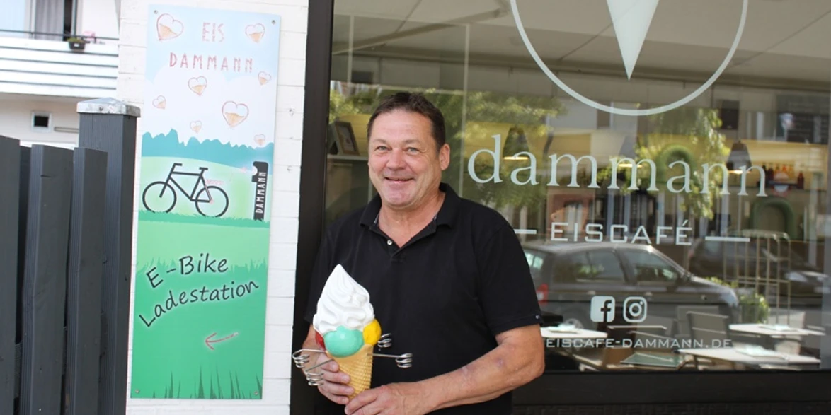 E-Ladesäule am Eiscafé Dammann Ein lächelnder Mann hält ein Eis vor dem Eingang eines modernen Eiscafés mit E-Bike-Ladestation.