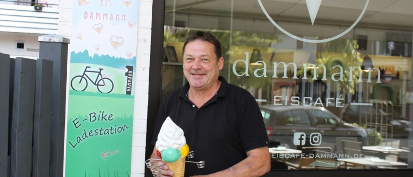 E-Ladesäule am Eiscafé Dammann Ein lächelnder Mann hält ein Eis vor dem Eingang eines modernen Eiscafés mit E-Bike-Ladestation.