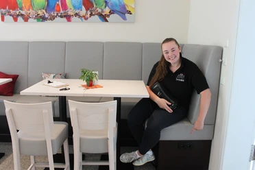 E-Bike-Ladesteckdosen im Cafe & Konditorei Tortenliebe Lächelnde Frau zeigt auf Steckdosen unter grauer Bank in modernem Café mit bunter Wanddekoration.