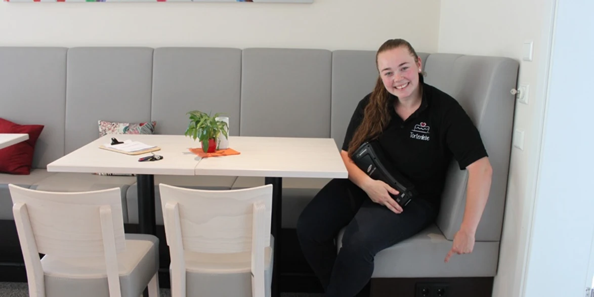 E-Bike-Ladesteckdosen im Cafe & Konditorei Tortenliebe Lächelnde Frau zeigt auf Steckdosen unter grauer Bank in modernem Café mit bunter Wanddekoration.