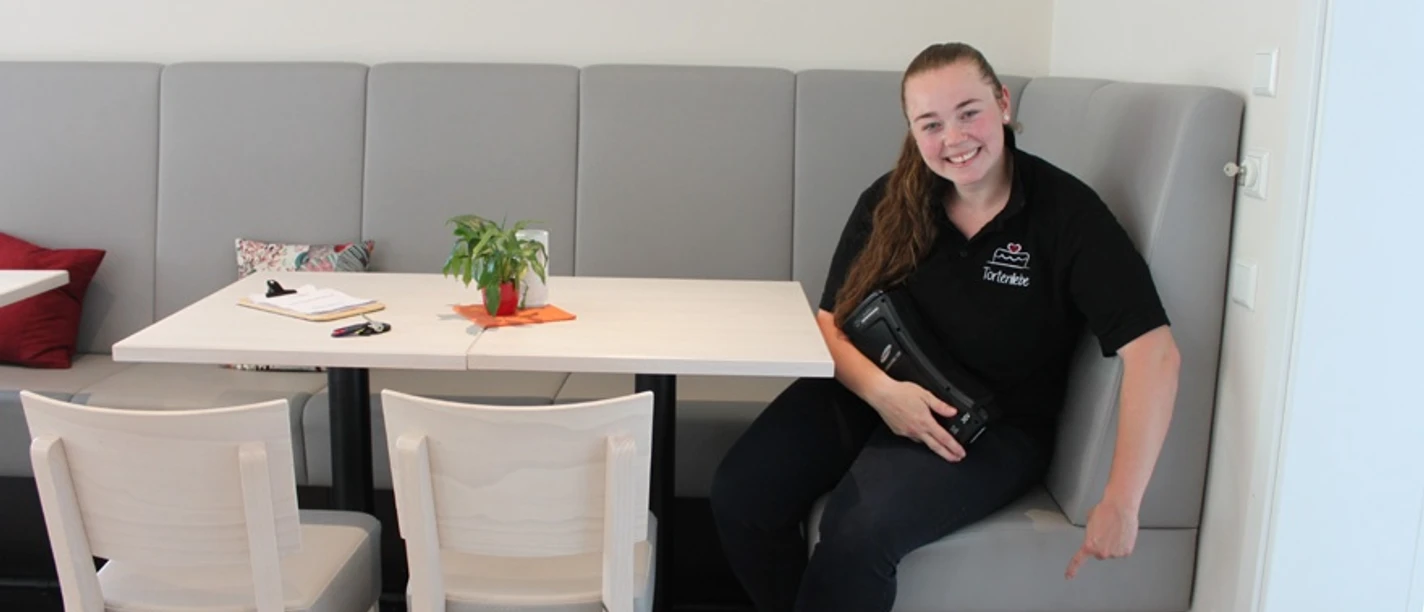 E-Bike-Ladesteckdosen im Cafe & Konditorei Tortenliebe Lächelnde Frau zeigt auf Steckdosen unter grauer Bank in modernem Café mit bunter Wanddekoration.