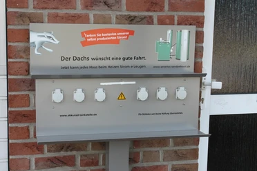 E-Bike-Ladesteckdosen, Andreas Fölling - Bad + Heizung Barrierefreie E-Tankstelle mit sechs weißen Steckdosen und einem Hinweisschild auf Backsteinwand.