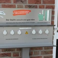 E-Bike-Ladesteckdosen, Andreas Fölling - Bad + Heizung Barrierefreie E-Tankstelle mit sechs weißen Steckdosen und einem Hinweisschild auf Backsteinwand.
