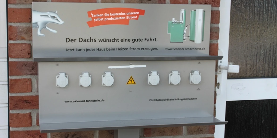 E-Bike-Ladesteckdosen, Andreas Fölling - Bad + Heizung Barrierefreie E-Tankstelle mit sechs weißen Steckdosen und einem Hinweisschild auf Backsteinwand.